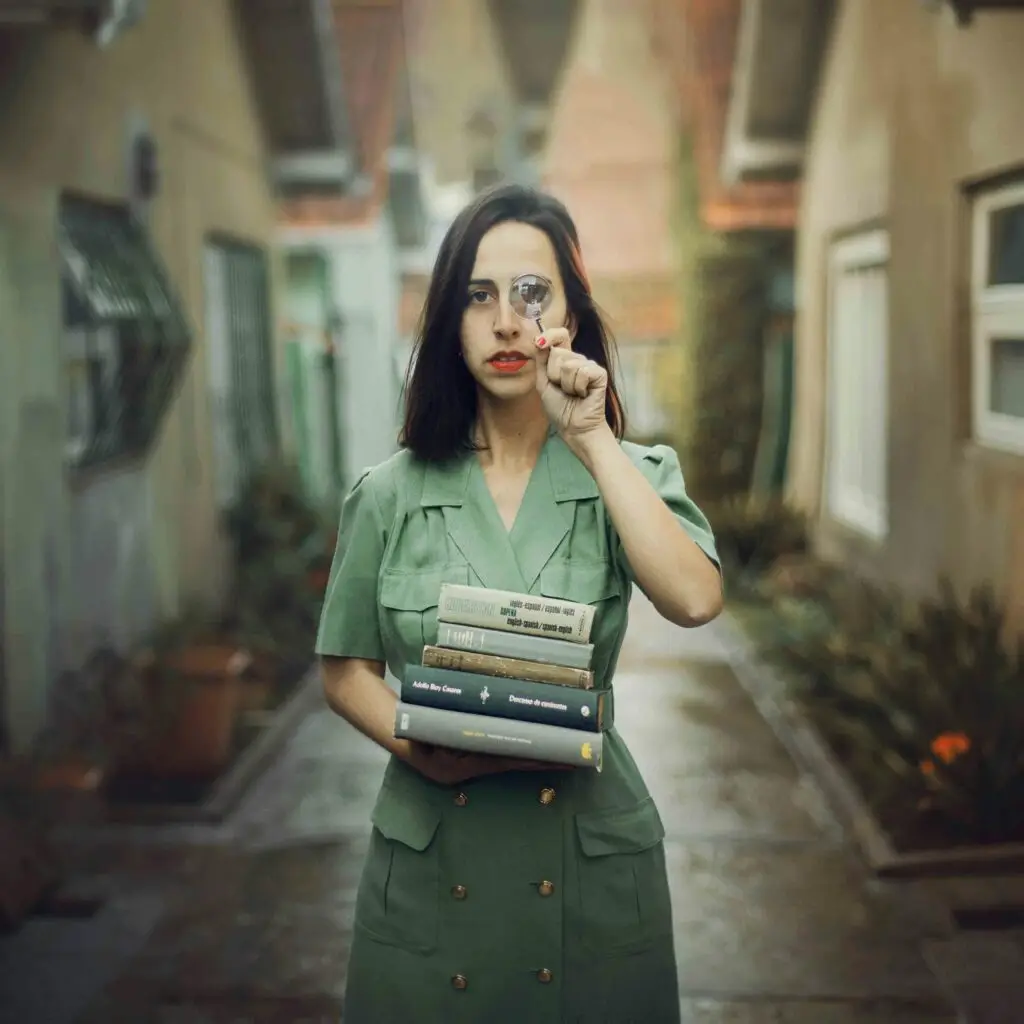 A woman with long hair in green dress holds books and magnifying glass standing in an alley.