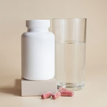 White bottle with red capsules and a glass of water on a beige background.